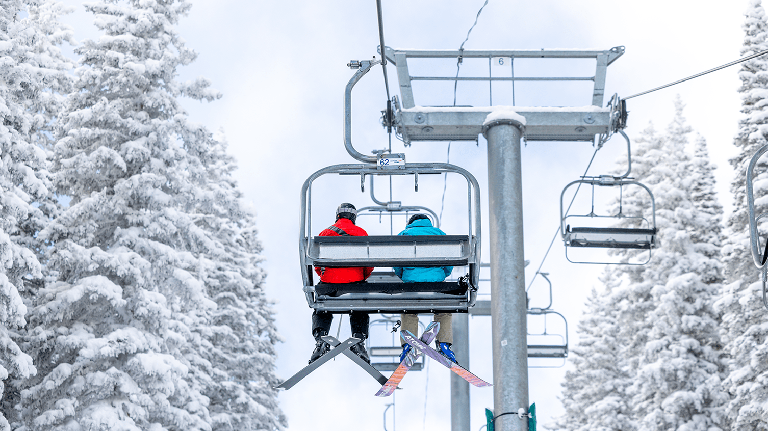 Two skiers sit on a chair lift, one in a blue coat, one in a red, Aspen Snowmass Pro coat. It is a bluebird day and the trees are coated in fresh snow