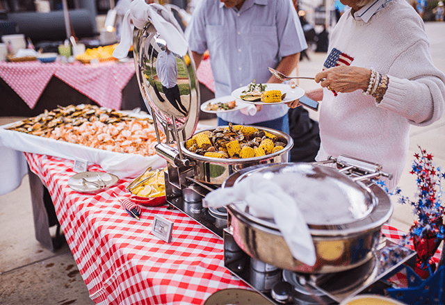 People serve themselves at a fancy buffet style food line up, corn and clams in a silver warmer, shrimp cocktail and festive gingham table cloth below