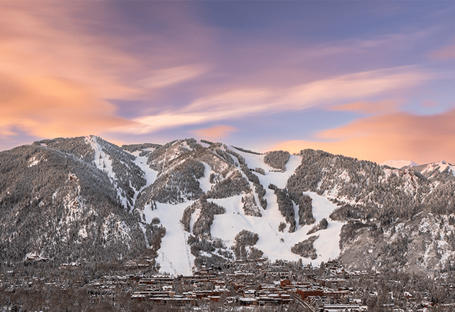 Long view of Aspen Mountain, at sunset, pink, orange and blue skies over white snowy runs, overlooking the town of Aspen