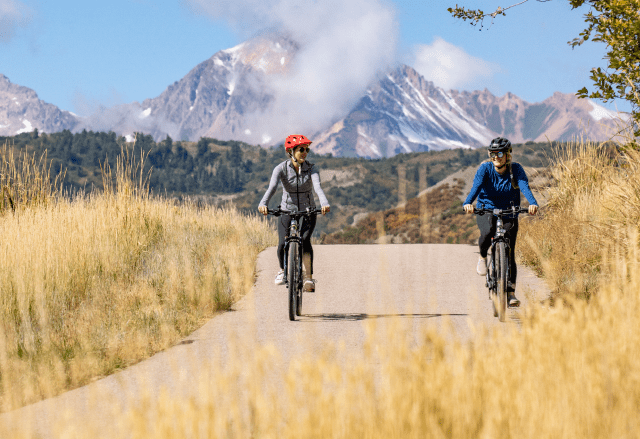 Couple rides in the late summer along a dirt road as snow starts to cap the peaks of Aspen Mountain