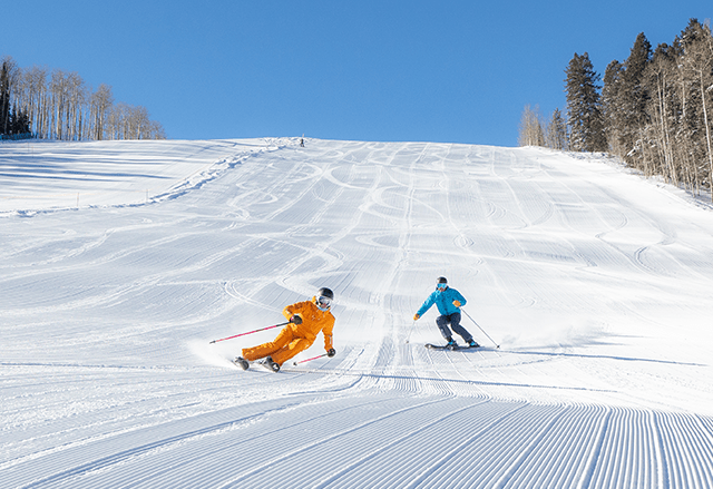 Skiers cruise down groomed run on a bluebird spring day at Aspen Snowmass