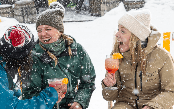three woman laugh as they hold drinks and snow falls atop their hats and ski jackets at Aspen Snowmass