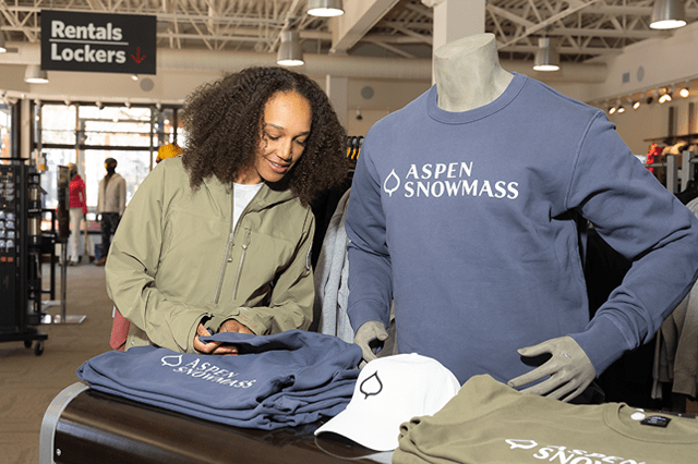 Guest looks through pile of blue gray aspen snowmass branded long sleeve t's at Four Mountain Sports
