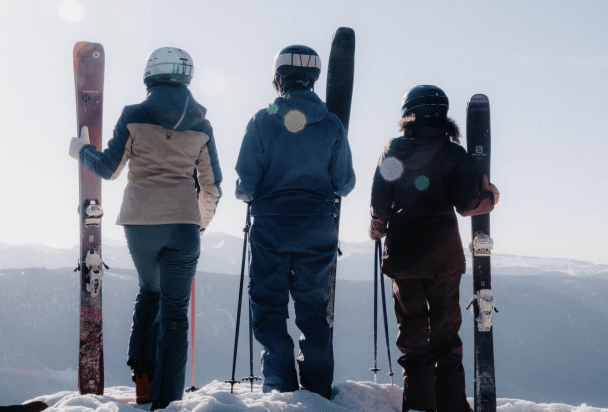 three people stand atop a mountain and look out, holding their skies