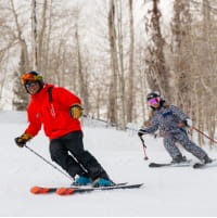 Kid skiing behind a ski pro in a red jacket near trees