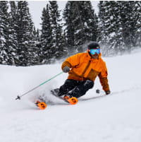 Skier in orange outfit skiing through powder in front of trees