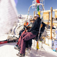 Three woman sit on the single chair lift chair atop aspen highland's bowls peak
