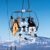 three snowboarders give the camera thumbs up as they ride over the camera on a chair lift