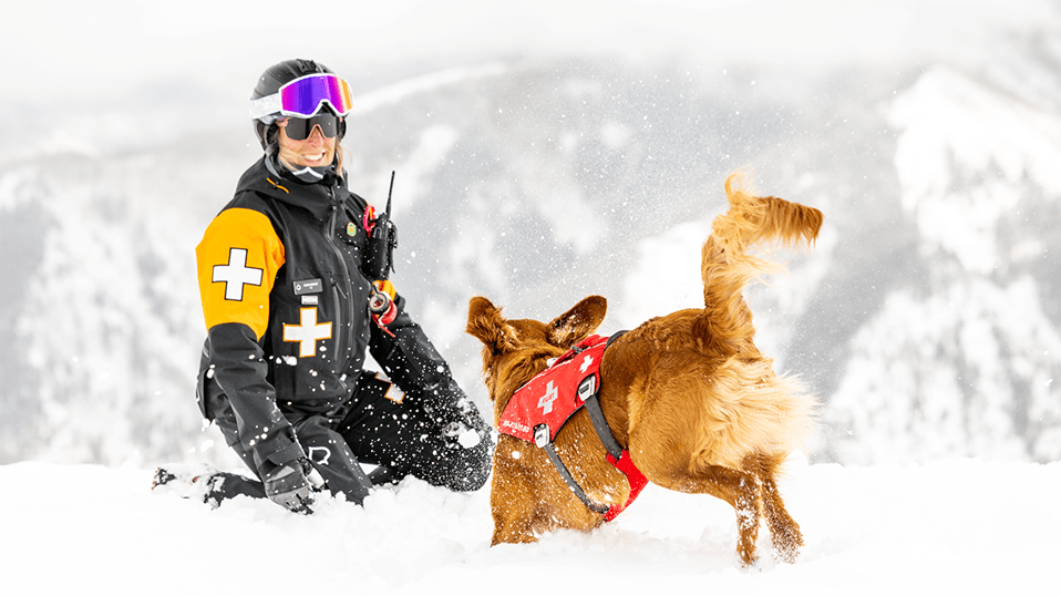 Female patroller sits in the snow as her golden retriever patrol dog bounds excitedly through the snow at her