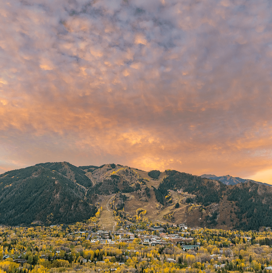 Fall shot of the town of Aspen and the mountain above, colored with green, yellow and orange aspen trees, with a pink and orange sunset over the fall town