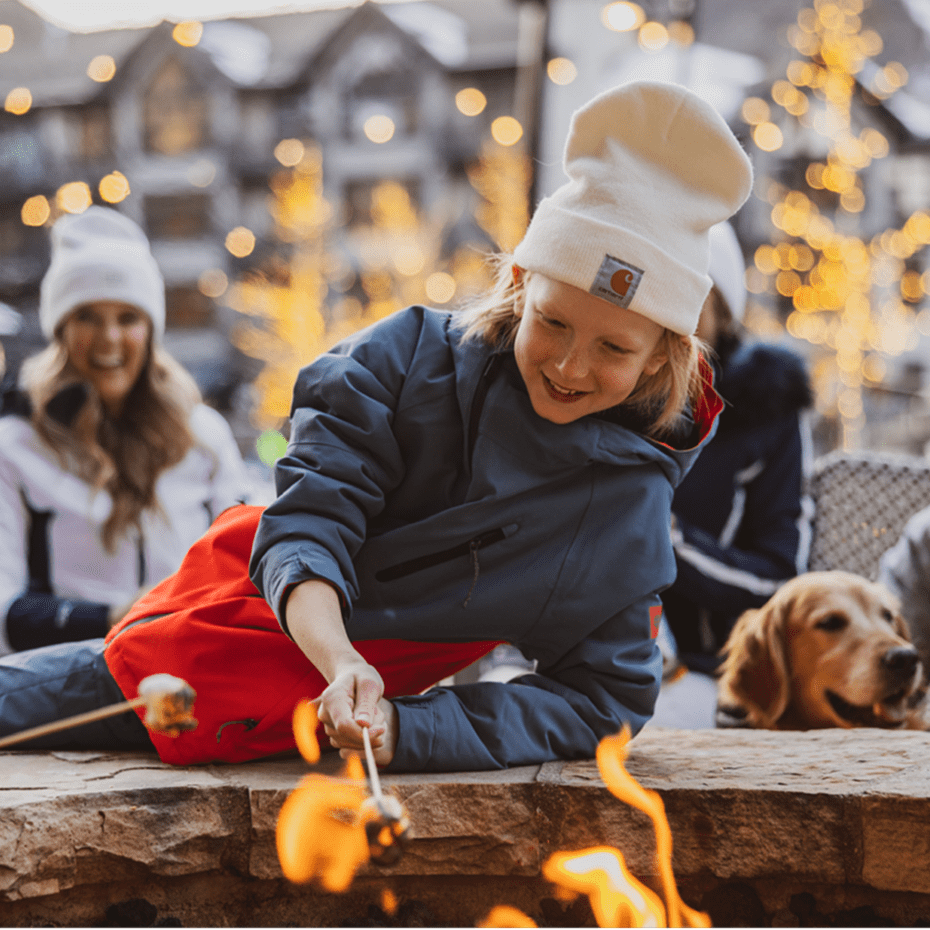 Little boy with white hat leans over the fireplace at Snowmass as he roasts his marshmallow over a firepit with his family