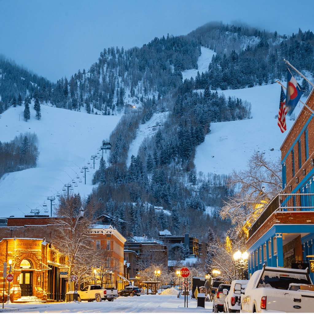 Aspen Snowmass at dusk, bright lights reflect off the snowy street, ski hill behind town