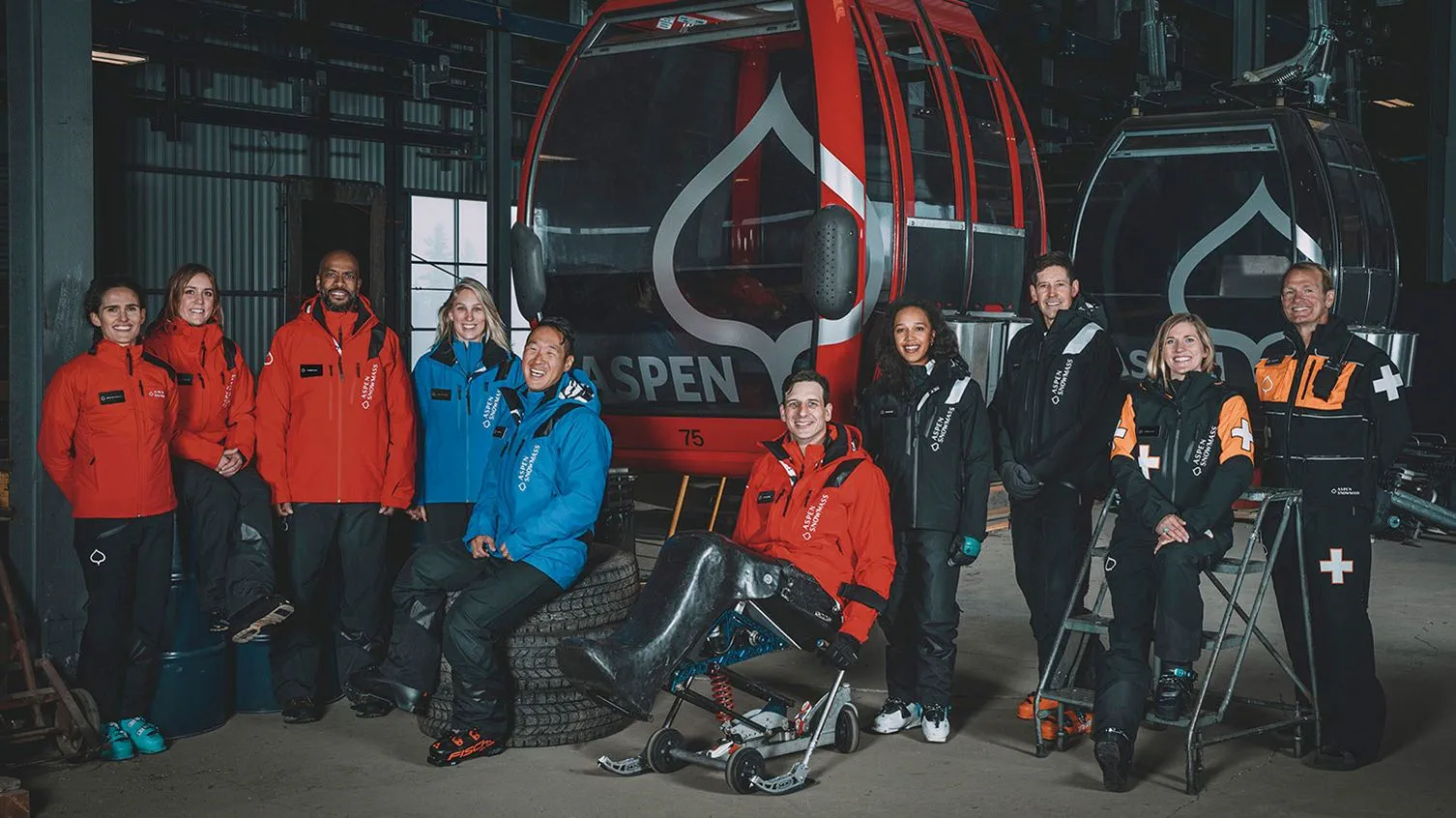 Nine people in winter clothing, including ski staff, stand beside an Aspen Snowmass red gondola indoors.