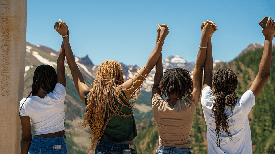 Four Women hold their hands victoriously up as they look out at Pyramid Peak and Aspen Snowmass