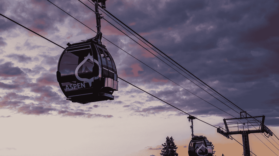 View of silver queen gondola from below, two black gondola cars on the cable, pink, purple and orange skies and clouds above, as the sun sets on Aspen Mountain