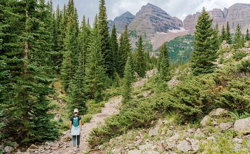 Woman hikes up a rocket trail towards the maroon bells, in the maroon bells wilderness, in the summer. Tall pine trees line the trail and the bells can be seen in the background.