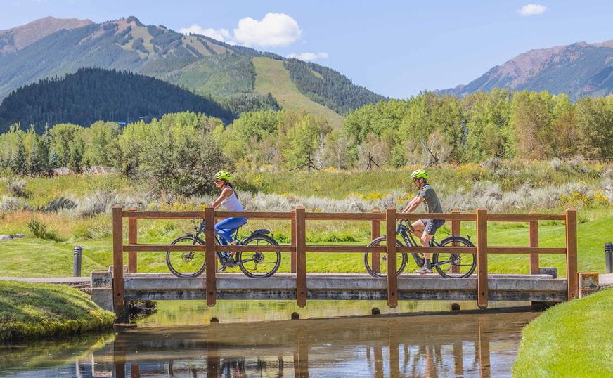 A couple rides across a bridge over the river in Aspen on a summer day, with Aspen Mountain in the background