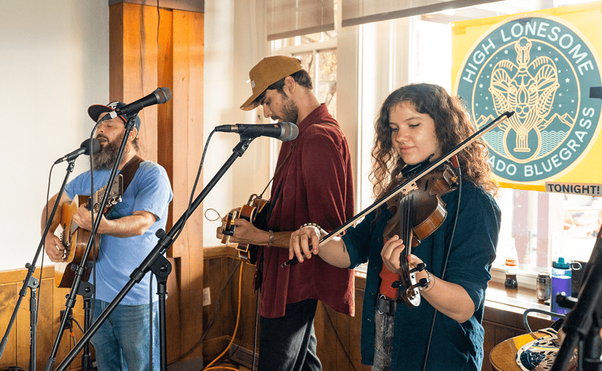 Three musicians play instruments at the sundeck for a crowd of people, a woman plays the violin and one man plays the mandolin and one plays the guitar