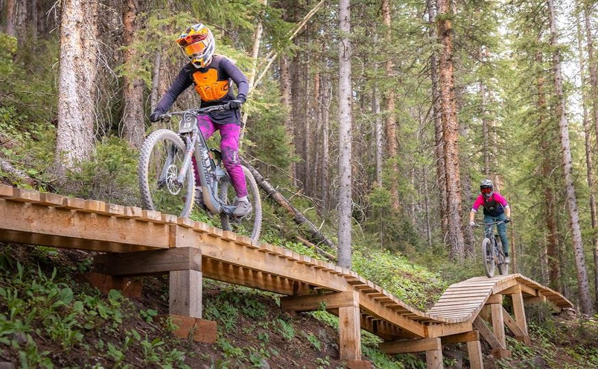 Two mountain bikers ride single file on a wooden board feature at Snowmass bike park, dressed head to toe in downhill gear