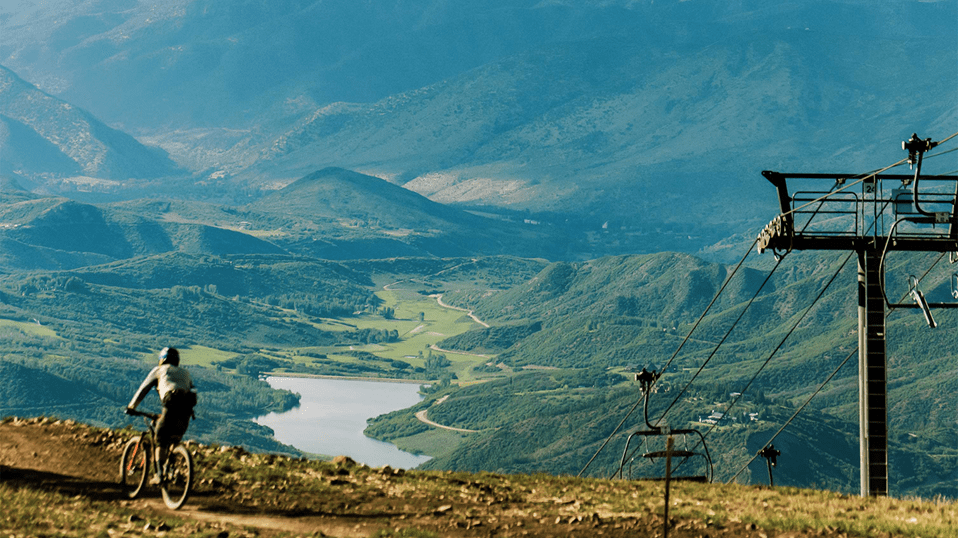 Downhill Mountain Biker cruises down a single track line at the snowmass bike park. The valley spreads out below, with green rolling hills and a beautiful blue lake. Chairlift poles rise up the mountain.