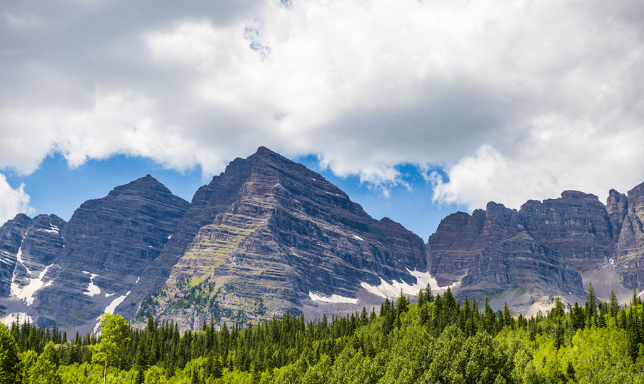 Maroon Bells over a green treeline, blue skies atop the mountains as clouds form above the mountains