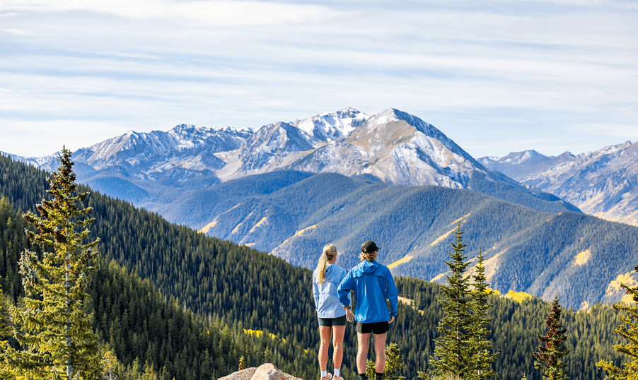 A couple stands on the lookout of Ajax, in early summer, green hills with snow still on the peaks of the mountains behind
