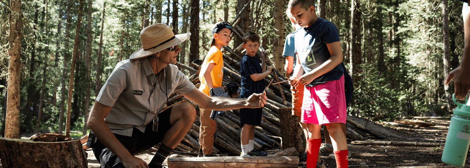 camp counselor hands a child something at Camp Aspen Snowmass day camp, in tall pine trees in the summer