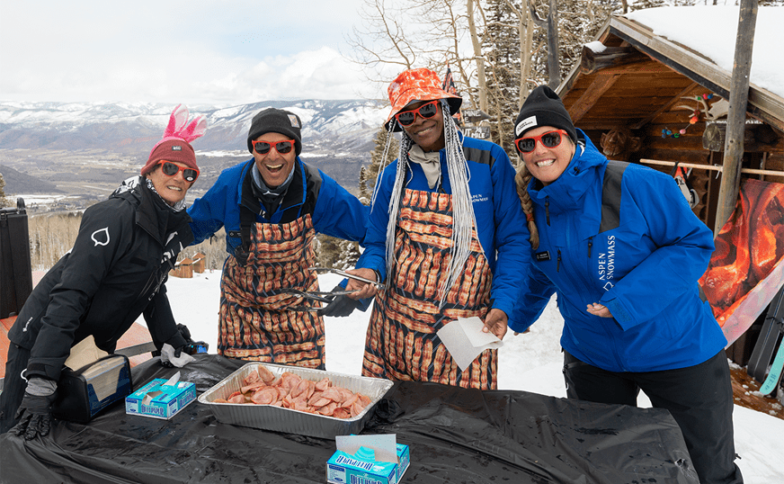 Four Buttermilk Employees smile with their arms around each other, as they stand at a table atop the mountain and hand out pieces of bacon for closing day