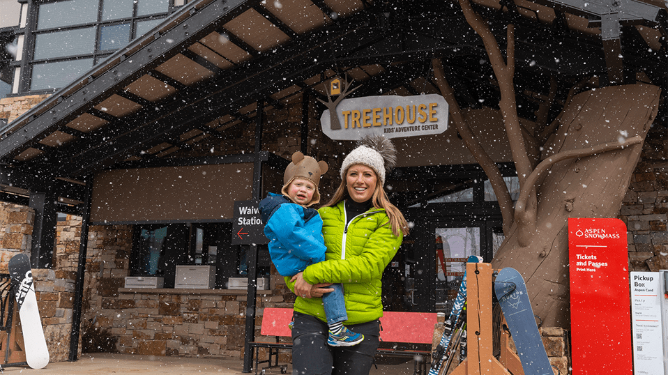 Mother dressed in a green coat holds her toddler in his blue coat and bear hat outside of the Treehouse at Aspen Snowmass
