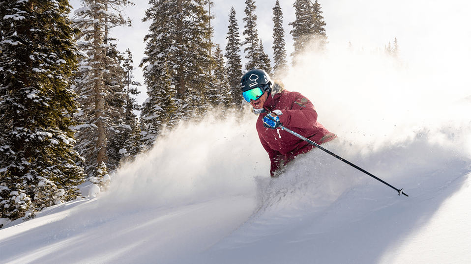 A skier in a red coat smiles in hip deep powder on a sunny power day, at aspen Snowmass
