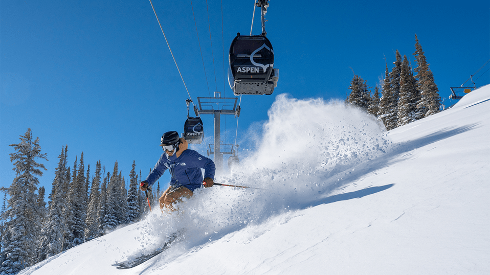 Skier carves under the Aspen Gondola, in deep powder, on a bluebird day