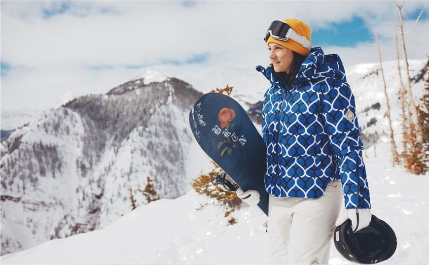 Woman in aspen collection jacket walks with her snowboard at her side on the mountain, in front of stunning mountain scape