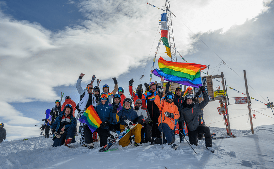 Large group of people stand atop the Aspen Highlands bowl, with large pride flags flying above them along side the bowl prayer flags