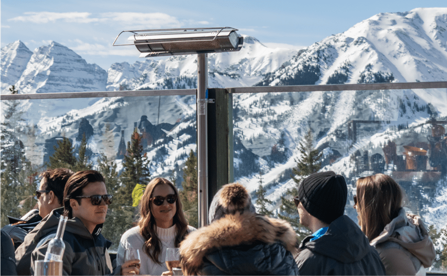 Group sits outside at Cloud nine, heat lamps warm them as snowy maroon bells stand tall behind them