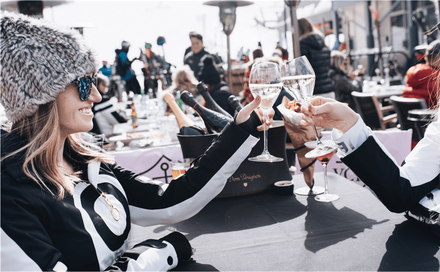 Two women in black and white ski sweaters cheers their wine glasses on a snowy day at the Base of Aspen Mountain, at Ajax Tavern