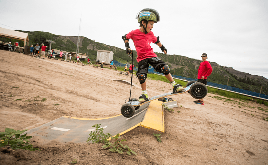Young boy rides a mountain board during Aspen Snowmass's summer camp, instructor coaches him from afar