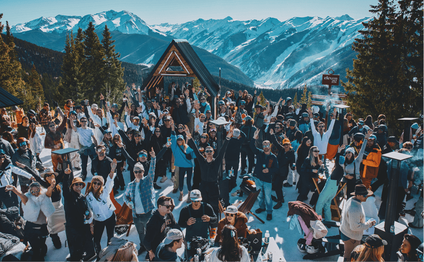 Crowd of people cheer at the top of Aspen Mountain at the Eleven212 apres spot, DJ plays and snowy peaks in the background
