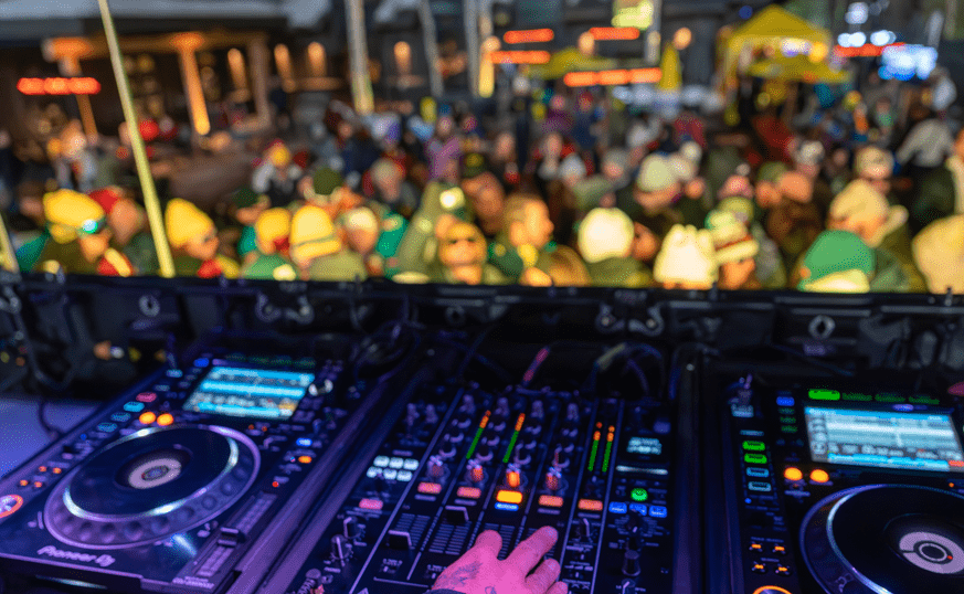 close up of DJ sound board, DJ's hand, and a crowd of people cheering in front of the stage