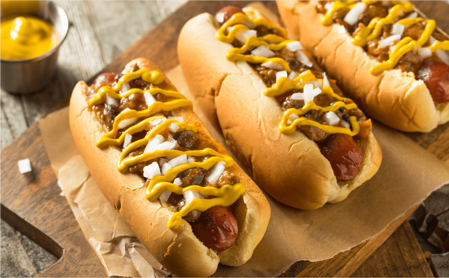 Close up image of three hotdogs with mustard and onions stacked on parchment paper, on a wodden table at Snowmass Ski resort
