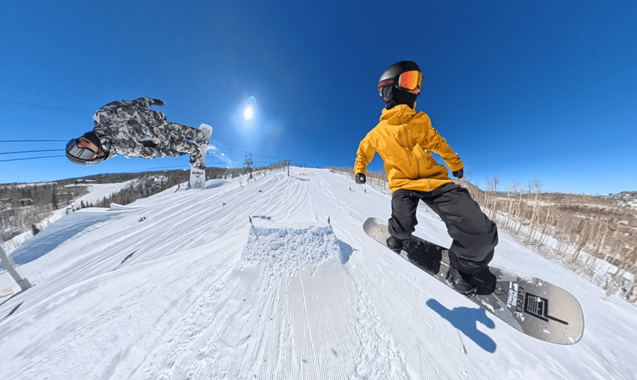 Two athletes snowboard down the terrain park on a bluebird day at Snowmass with their Insta360