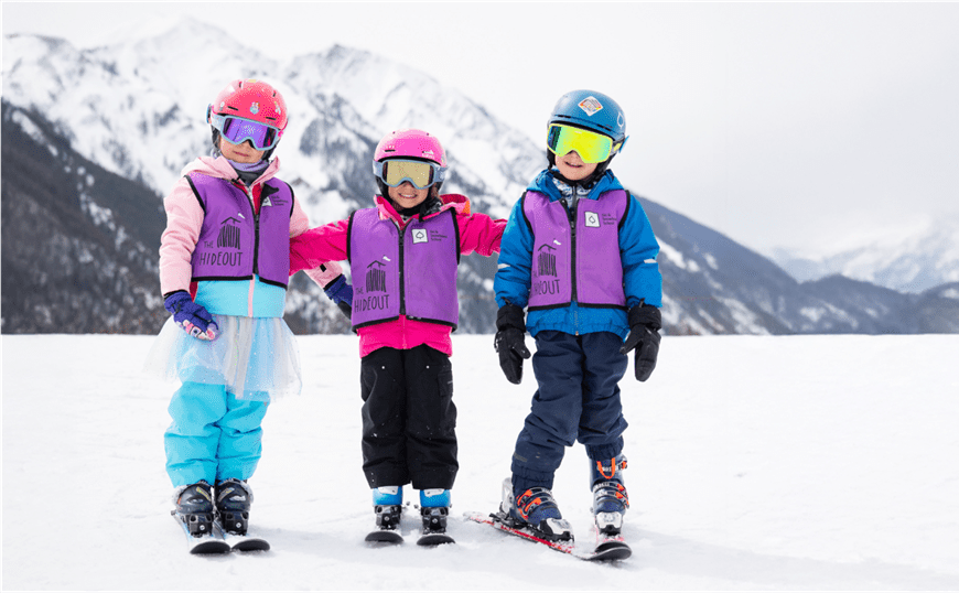 Three kids smile at the camera atop of Buttermilk mountain, they are dressed in their lesson gear 
