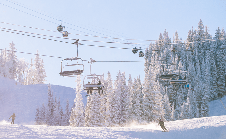 Aspen mountain on a glittery snowy morning, people ski under the aspen ski lifts as the sun starts to rise over the mountains