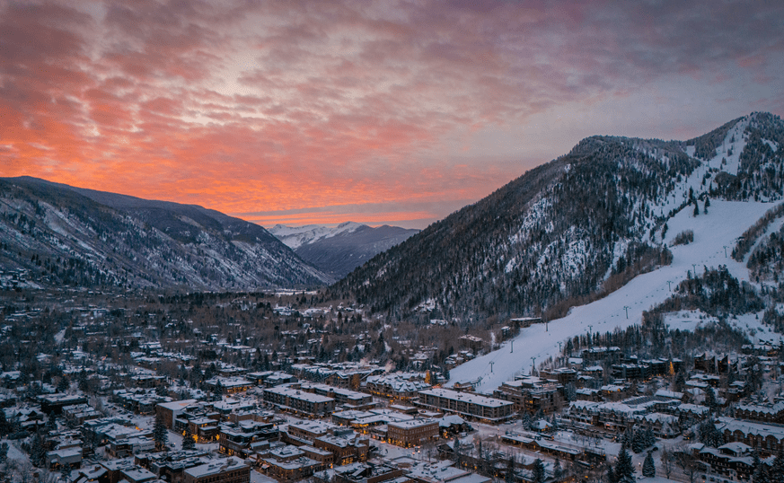 The town of aspen at sunset, pink and orange skies over a snowy town, the runs of Aspen Mountain to the right