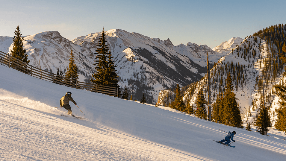 Two skiers carve down a groomed run at snowmass, as golden  sunlight lights up the mountians behind