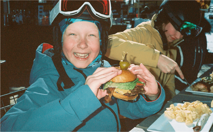 Little boy in ski gear smiles as he holds a huge cheeseburger at Snowmass ski resort
