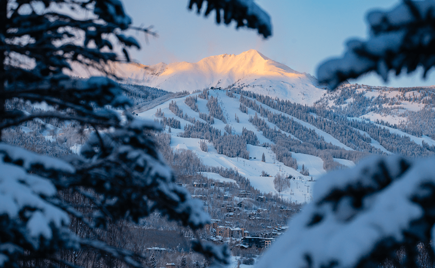 View of snowmass ski resort through snowy trees on a winter evening, sun sets over ski runs