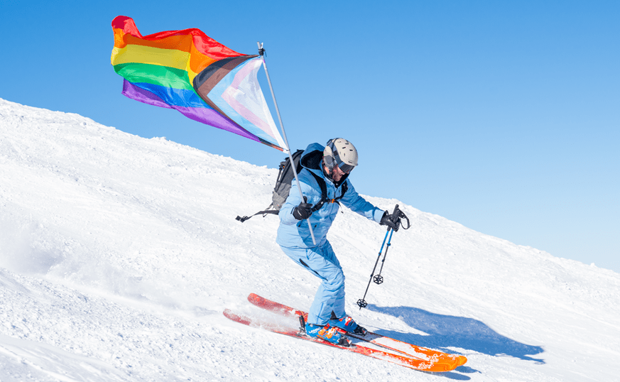 Skier in a blue ski kit carves down a groomed run, blue skies behind, with the LGBTQ+ flag streaming behind them