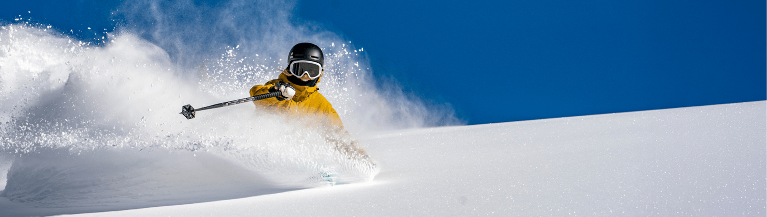 Shot of skier head on as they carve down the mountain, in a yellow ski jacket, in deep powder and under deep blue skies on Aspen Ski Resort