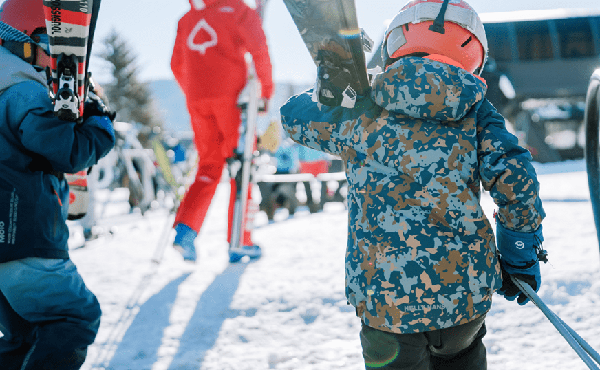 two small children walk behind ski instructor with their skies in hand