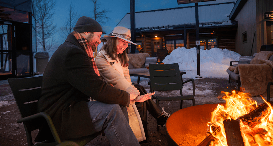 A couple warms their hands over the fire at the cabin at snowcat dinners